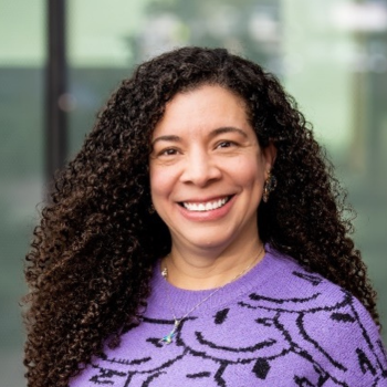 Woman with long curly hair smiling, wearing purple top, indoor background